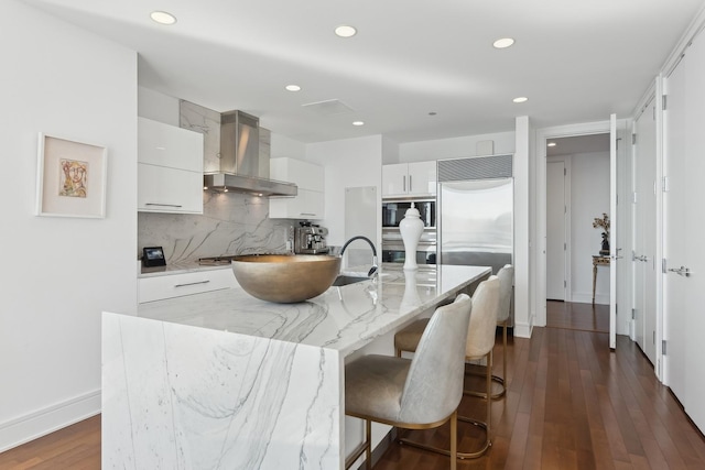 kitchen with a breakfast bar area, dark wood-style floors, built in appliances, wall chimney exhaust hood, and tasteful backsplash