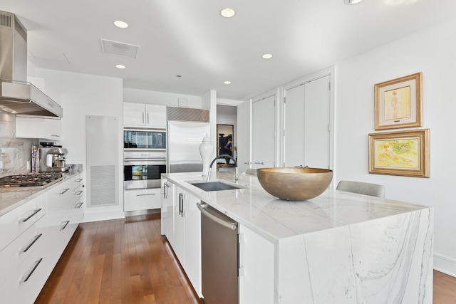 kitchen with dark wood-style floors, a sink, built in appliances, wall chimney range hood, and tasteful backsplash