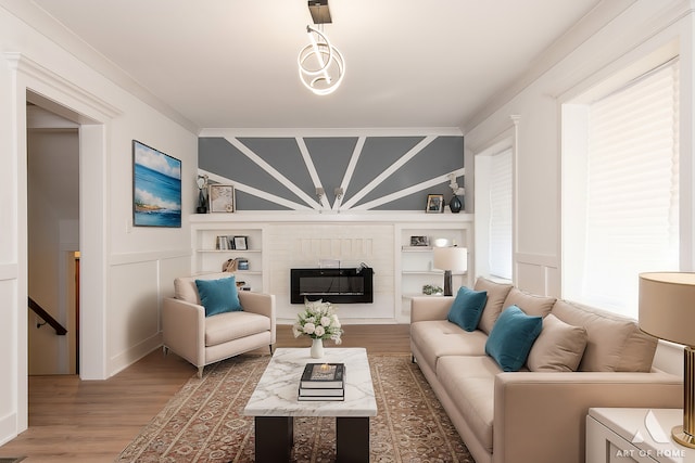 living area with a decorative wall, built in features, ornamental molding, wood finished floors, and a glass covered fireplace