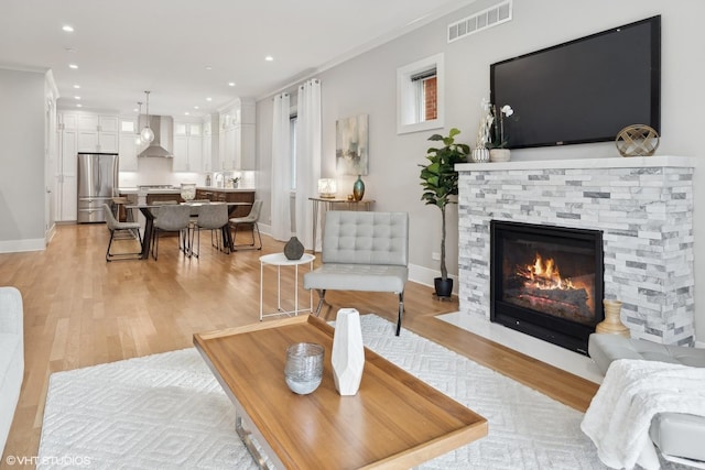 living area featuring light wood finished floors, visible vents, crown molding, baseboards, and a stone fireplace