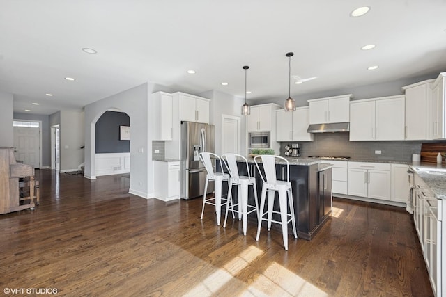 kitchen with dark wood-style flooring, arched walkways, stainless steel appliances, under cabinet range hood, and a center island