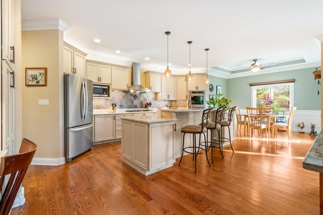 kitchen featuring a kitchen bar, wall chimney range hood, appliances with stainless steel finishes, light countertops, and ceiling fan