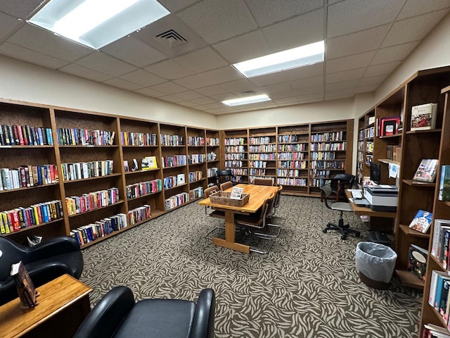 carpeted office featuring visible vents, a paneled ceiling, and wall of books