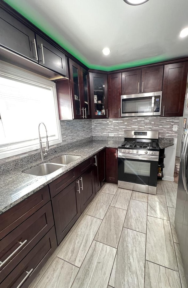 kitchen featuring backsplash, glass insert cabinets, appliances with stainless steel finishes, stone countertops, and a sink