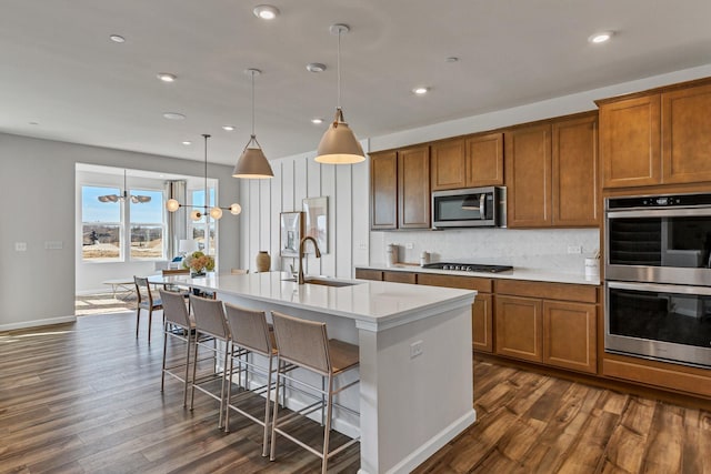 kitchen featuring an island with sink, a sink, tasteful backsplash, appliances with stainless steel finishes, and brown cabinetry