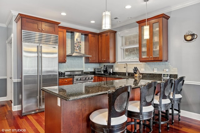 kitchen featuring dark stone countertops, stainless steel appliances, a peninsula, wall chimney range hood, and glass insert cabinets