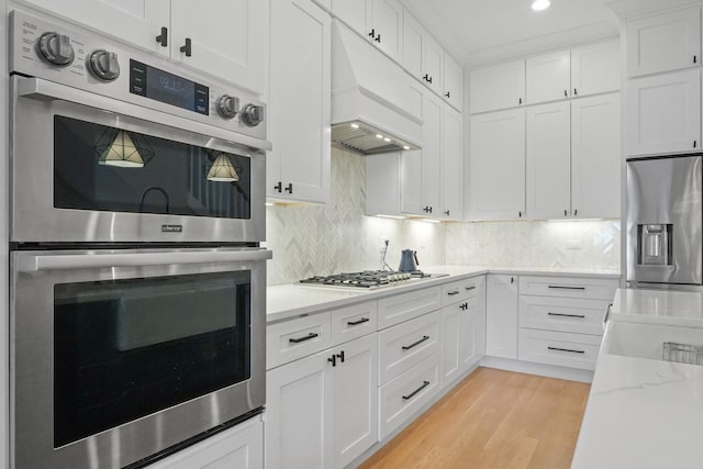 kitchen featuring premium range hood, decorative backsplash, stainless steel appliances, light wood-style floors, and white cabinetry