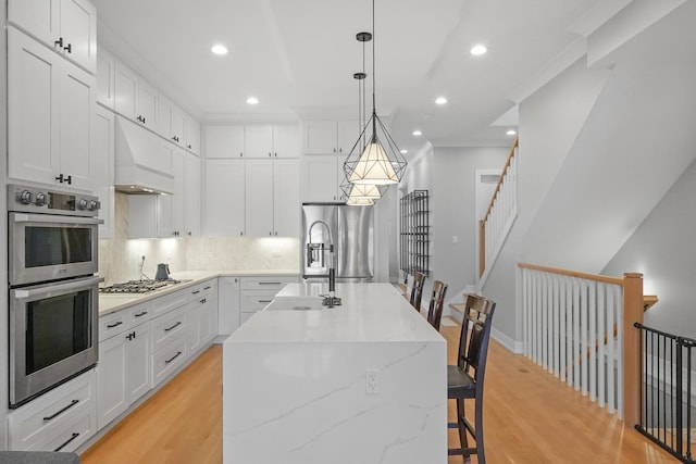 kitchen with light wood-type flooring, custom exhaust hood, appliances with stainless steel finishes, and an island with sink