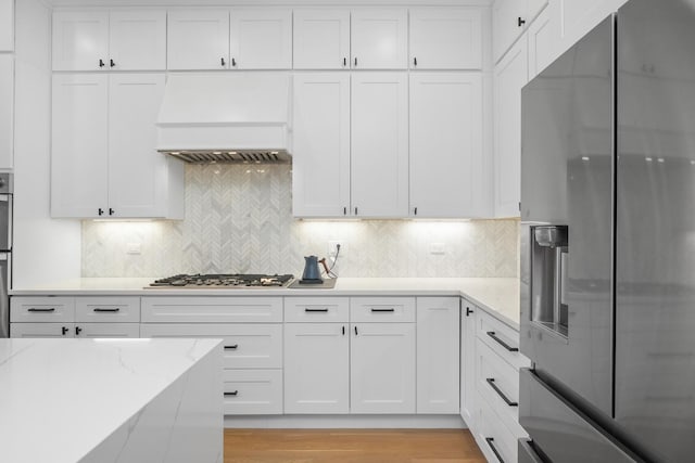 kitchen with backsplash, light stone countertops, custom range hood, white cabinets, and stainless steel appliances