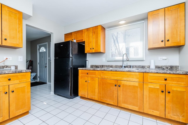 kitchen featuring a sink, dark stone countertops, freestanding refrigerator, brown cabinetry, and light tile patterned floors