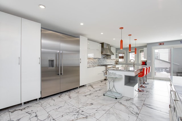 kitchen featuring stainless steel built in refrigerator, a sink, tasteful backsplash, wall chimney exhaust hood, and white cabinets