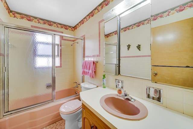 full bathroom with vanity, a wainscoted wall, bath / shower combo with glass door, tile walls, and toilet