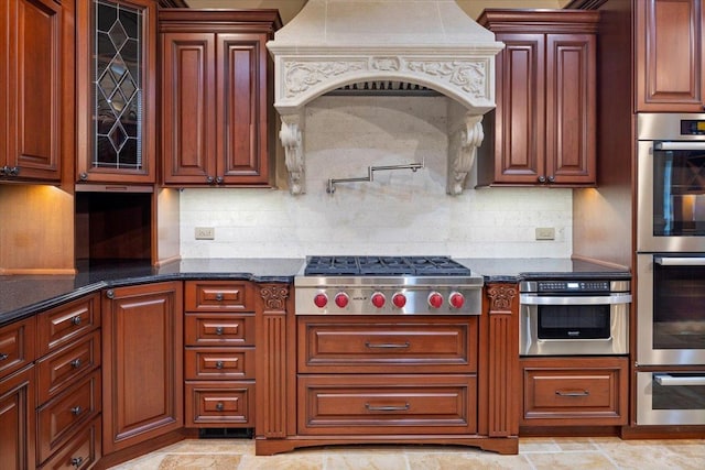 kitchen with dark stone countertops, custom exhaust hood, glass insert cabinets, appliances with stainless steel finishes, and tasteful backsplash