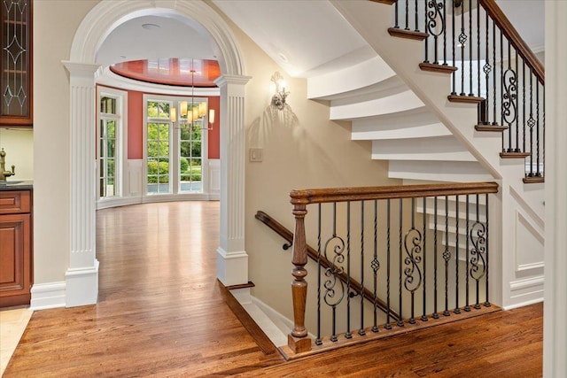 stairway featuring baseboards, a tray ceiling, decorative columns, an inviting chandelier, and wood finished floors