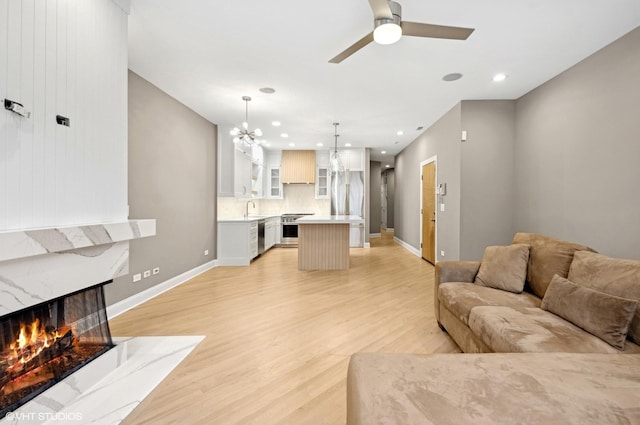 living room with baseboards, light wood-type flooring, a premium fireplace, recessed lighting, and ceiling fan with notable chandelier