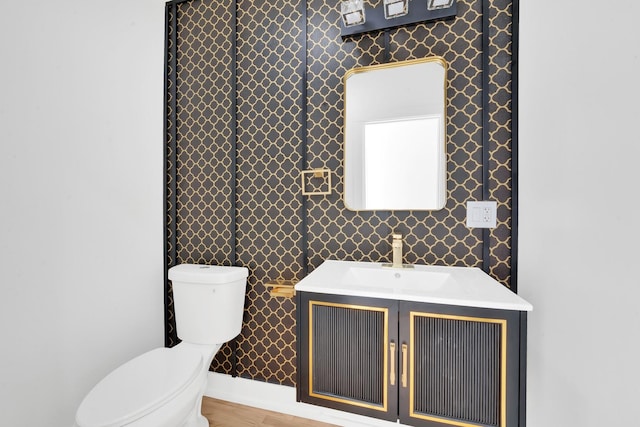 bathroom featuring vanity, wood finished floors, baseboards, wallpapered walls, and toilet