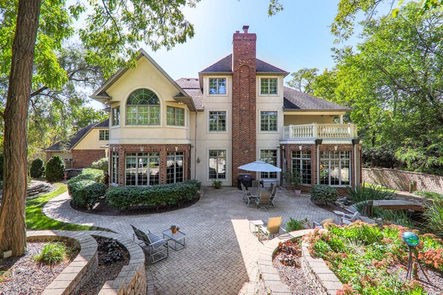 rear view of house with brick siding, stucco siding, a chimney, a balcony, and a patio area