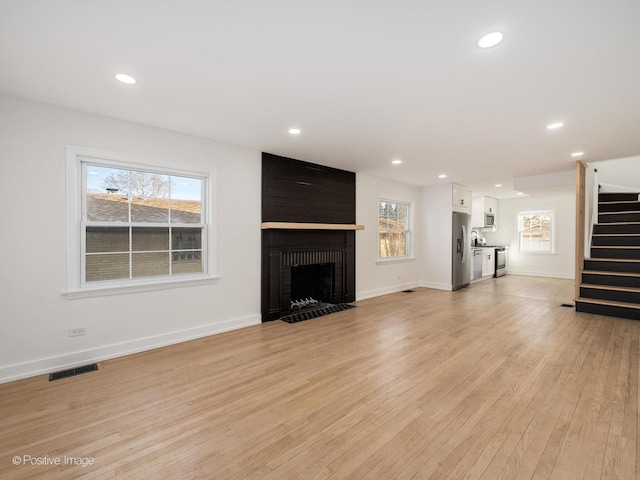 unfurnished living room featuring visible vents, light wood finished floors, a brick fireplace, stairs, and a healthy amount of sunlight