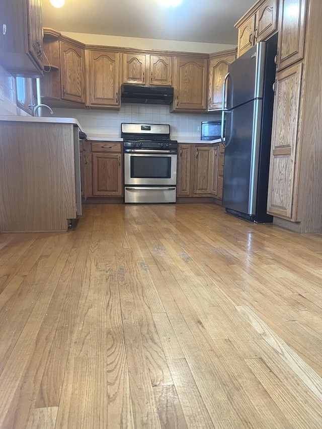 kitchen featuring backsplash, under cabinet range hood, light wood-type flooring, brown cabinetry, and stainless steel appliances