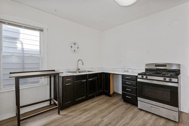 kitchen featuring light stone counters, dark cabinets, a sink, stainless steel gas range oven, and light wood-type flooring