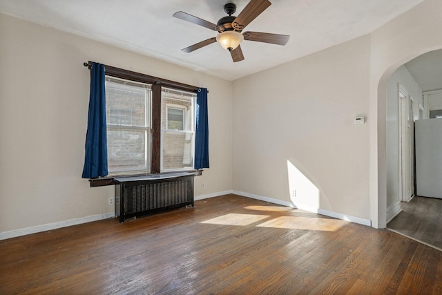 spare room with a ceiling fan, arched walkways, radiator, wood-type flooring, and baseboards