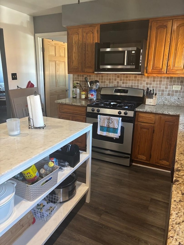 kitchen with dark wood-type flooring, backsplash, stainless steel appliances, brown cabinetry, and light stone countertops