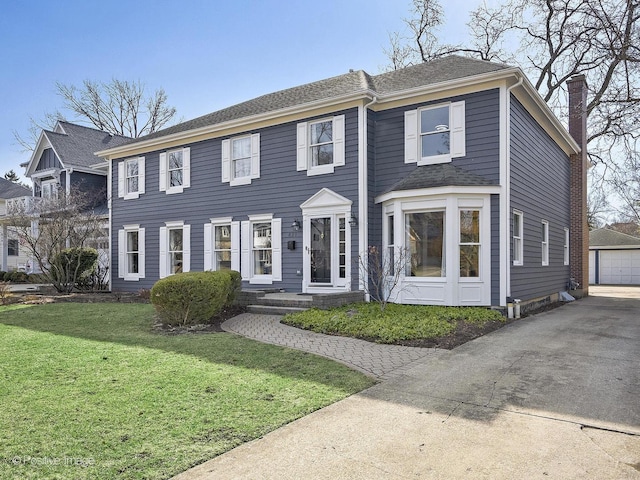 colonial-style house featuring an outbuilding, a shingled roof, a chimney, a front lawn, and a detached garage