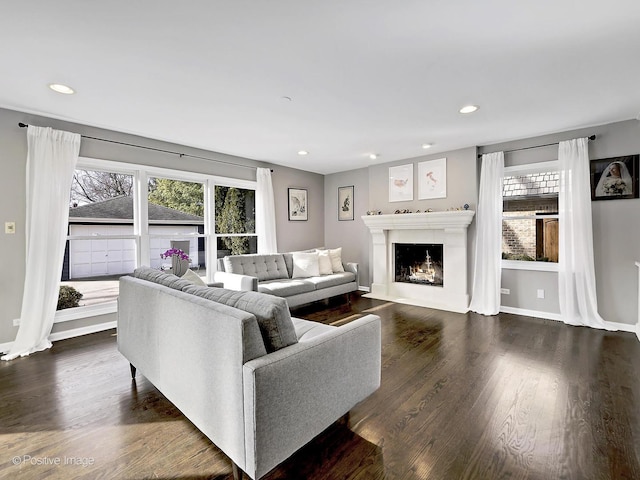 living room featuring recessed lighting, baseboards, dark wood-type flooring, and a warm lit fireplace