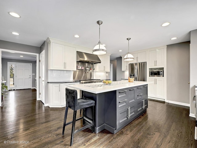 kitchen featuring dark wood-type flooring, wall chimney range hood, decorative light fixtures, white cabinets, and stainless steel appliances