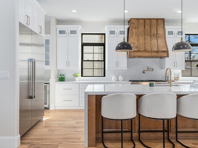 kitchen with premium range hood, glass insert cabinets, white cabinetry, light wood-type flooring, and stainless steel built in refrigerator