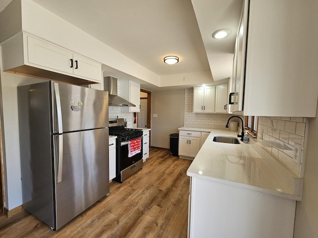 kitchen with wall chimney range hood, wood finished floors, white cabinets, stainless steel appliances, and a sink