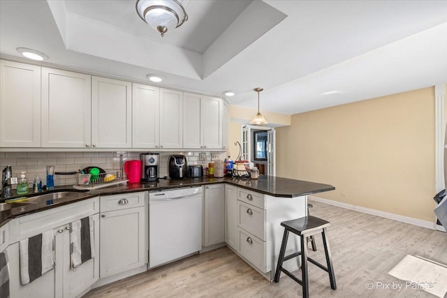 kitchen with light wood-type flooring, a tray ceiling, a peninsula, white dishwasher, and tasteful backsplash