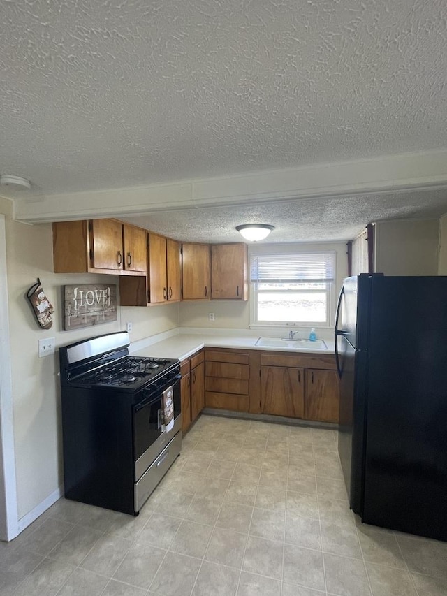 kitchen featuring stainless steel range with gas stovetop, freestanding refrigerator, a sink, light countertops, and brown cabinets