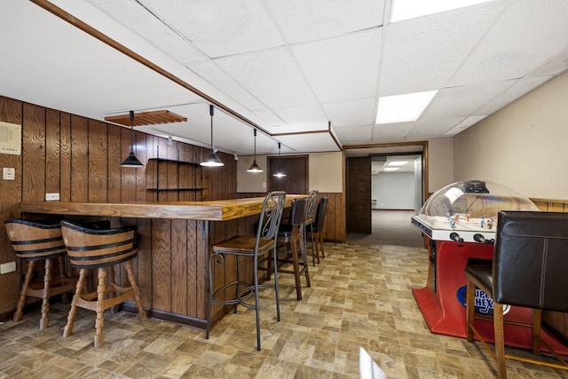 bar featuring decorative light fixtures, a drop ceiling, stone finish flooring, a bar, and wood walls