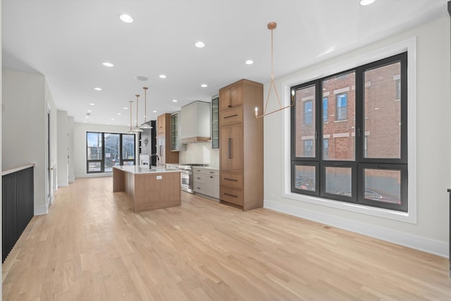 kitchen with wall chimney range hood, an island with sink, recessed lighting, light wood-style flooring, and stainless steel stove