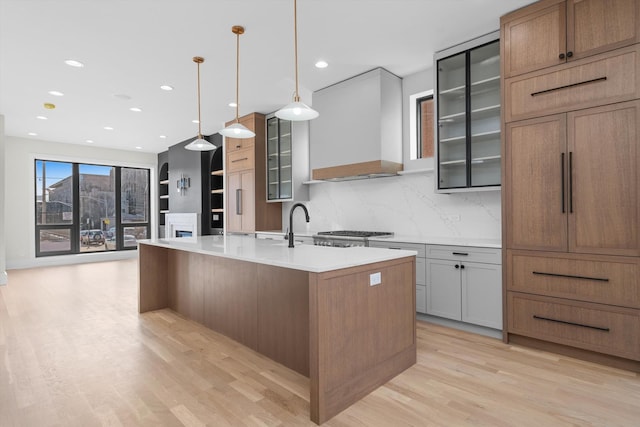 kitchen featuring a center island with sink, premium range hood, light wood finished floors, light countertops, and glass insert cabinets