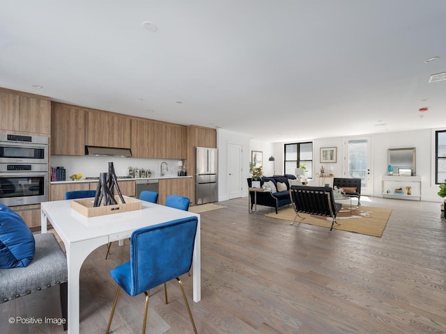 kitchen with brown cabinetry, light wood-type flooring, under cabinet range hood, appliances with stainless steel finishes, and open floor plan