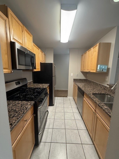 kitchen with light brown cabinetry, dark stone counters, light tile patterned floors, stainless steel appliances, and a sink