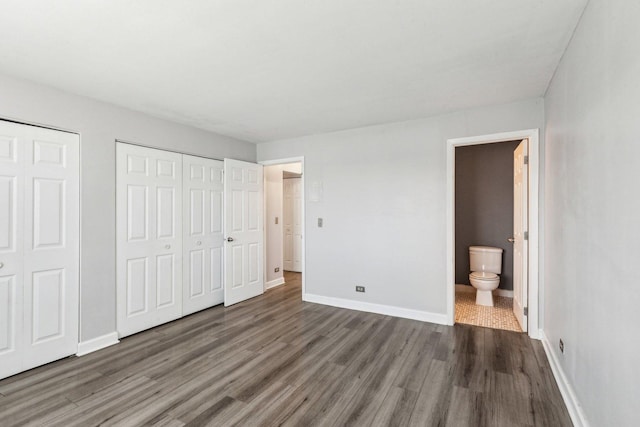 unfurnished bedroom with two closets, ensuite bathroom, baseboards, and wood finished floors