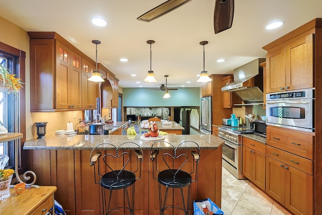 kitchen featuring wall chimney range hood, high quality appliances, light stone counters, a peninsula, and a ceiling fan