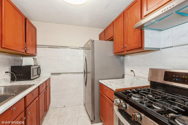 kitchen with under cabinet range hood, appliances with stainless steel finishes, brown cabinetry, marble finish floor, and a textured ceiling