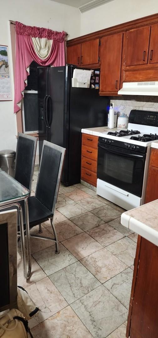 kitchen with backsplash, under cabinet range hood, light countertops, gas range oven, and black refrigerator with ice dispenser