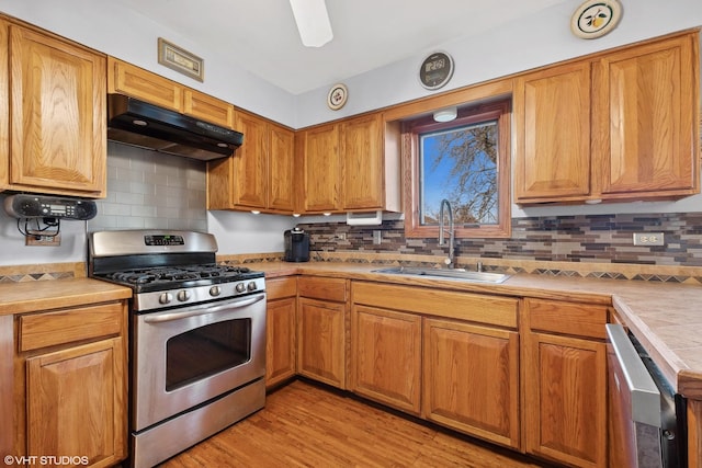 kitchen featuring under cabinet range hood, a sink, stainless steel appliances, brown cabinetry, and light countertops