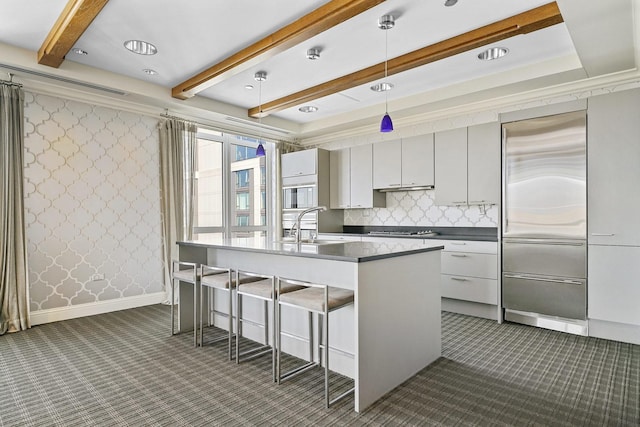 kitchen with wallpapered walls, beam ceiling, stainless steel built in fridge, a sink, and dark countertops