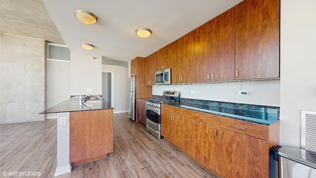 kitchen with brown cabinets, a peninsula, dark stone counters, appliances with stainless steel finishes, and light wood-type flooring