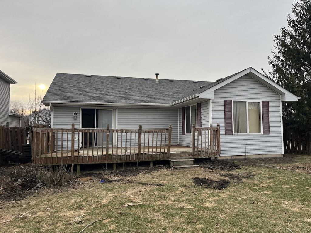 back of house at dusk featuring fence, a lawn, a deck, and a shingled roof