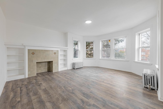 unfurnished living room with a tiled fireplace, radiator, built in shelves, and wood finished floors