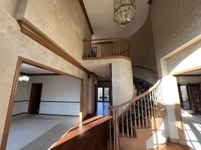 stairway featuring a high ceiling, an inviting chandelier, wainscoting, a decorative wall, and crown molding