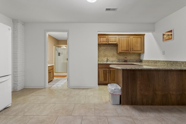 kitchen featuring dark stone countertops, visible vents, freestanding refrigerator, brown cabinets, and decorative backsplash