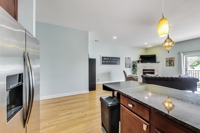 kitchen featuring stainless steel refrigerator with ice dispenser, dark stone counters, hanging light fixtures, light wood finished floors, and a fireplace